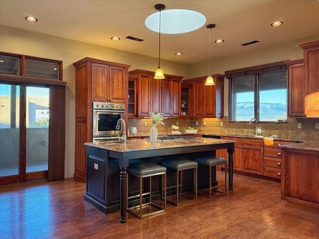 kitchen with dark hardwood / wood-style flooring, tasteful backsplash, light stone counters, decorative light fixtures, and an island with sink