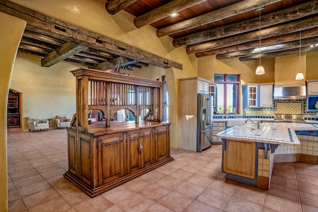 kitchen with wall chimney range hood, beam ceiling, stainless steel fridge with ice dispenser, tile counters, and an island with sink