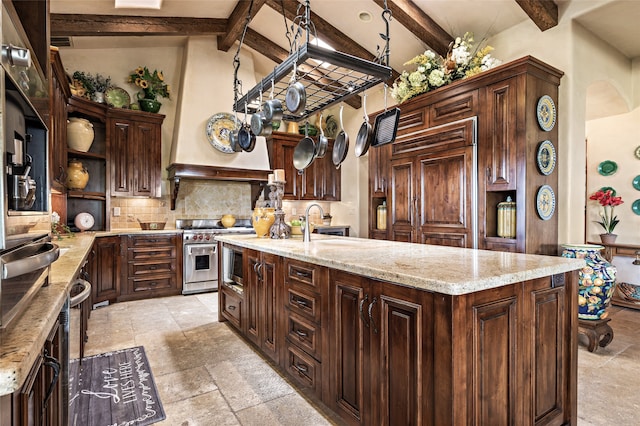 kitchen with dark brown cabinetry, light stone countertops, beamed ceiling, high quality appliances, and a center island with sink