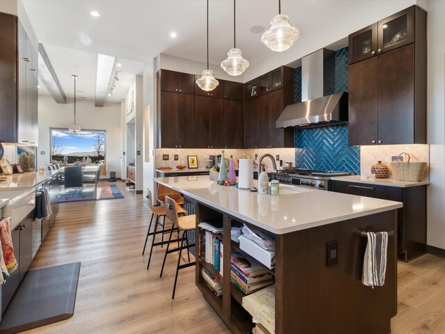 kitchen featuring wall chimney exhaust hood, light wood-type flooring, a kitchen island with sink, and decorative light fixtures