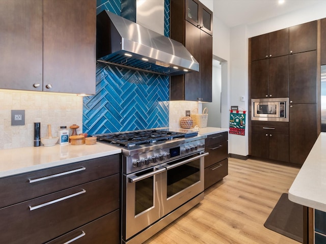 kitchen with wall chimney exhaust hood, light wood-type flooring, tasteful backsplash, dark brown cabinetry, and stainless steel appliances