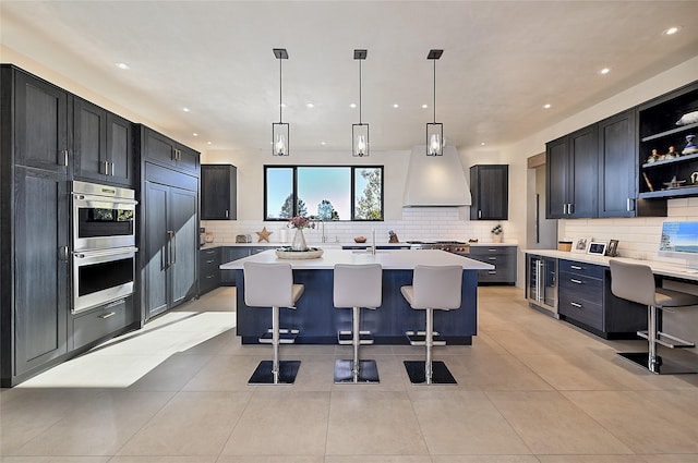 kitchen with custom exhaust hood, a kitchen island with sink, light tile patterned floors, hanging light fixtures, and a breakfast bar area