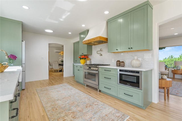 kitchen with decorative backsplash, premium range hood, stainless steel appliances, green cabinetry, and light hardwood / wood-style floors