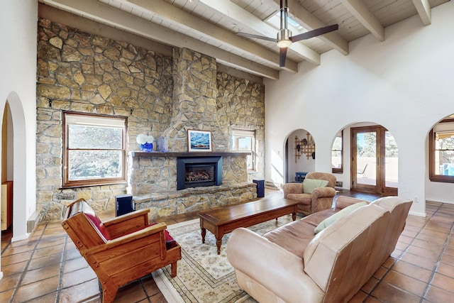 living room with a towering ceiling, beam ceiling, ceiling fan, wooden ceiling, and a stone fireplace