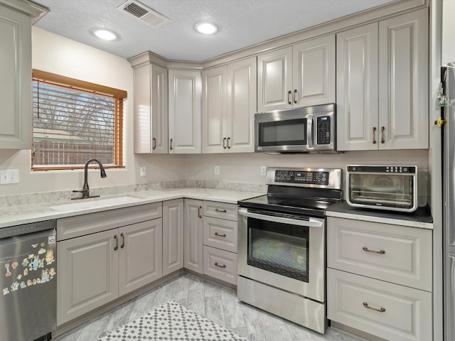 kitchen with gray cabinets, stainless steel appliances, light stone countertops, a textured ceiling, and sink