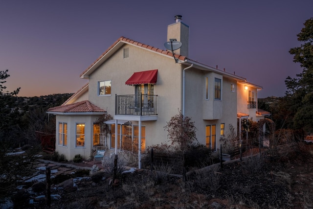 back of property at dusk featuring a tiled roof, a chimney, a balcony, and stucco siding