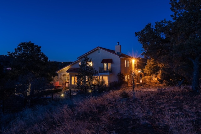 back of house at twilight featuring a chimney, a balcony, and stucco siding