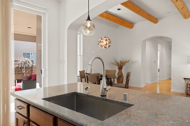 kitchen with light stone countertops, decorative light fixtures, light wood-type flooring, beam ceiling, and sink