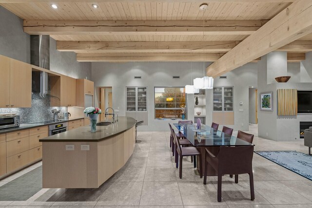 kitchen with oven, beamed ceiling, light brown cabinets, an island with sink, and hanging light fixtures