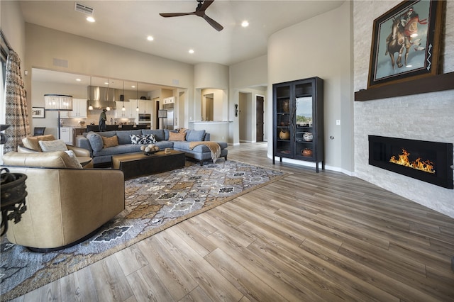 living room featuring hardwood / wood-style flooring, ceiling fan, a stone fireplace, and a high ceiling