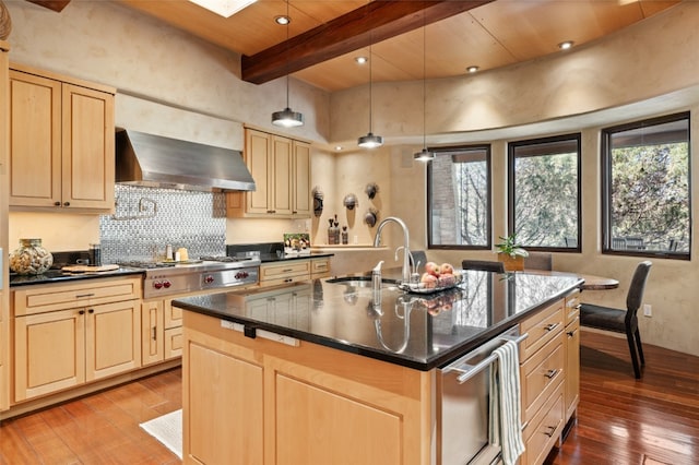 kitchen featuring decorative light fixtures, wall chimney range hood, sink, an island with sink, and light brown cabinets