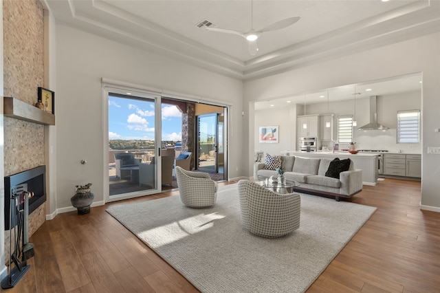 living room featuring ceiling fan, wood-type flooring, a raised ceiling, and a tiled fireplace