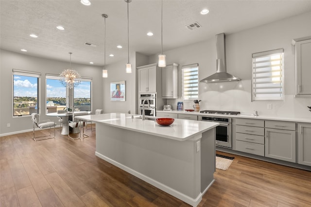 kitchen with decorative light fixtures, a center island with sink, light hardwood / wood-style flooring, wall chimney exhaust hood, and a chandelier