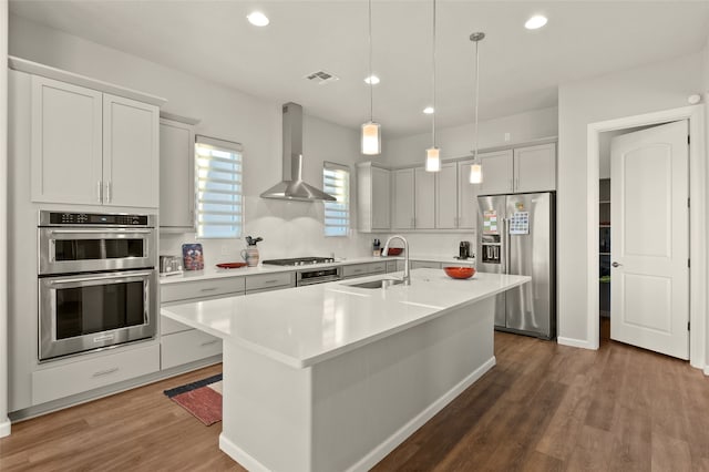 kitchen with pendant lighting, appliances with stainless steel finishes, wall chimney range hood, sink, and a center island with sink