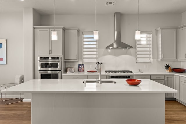 kitchen featuring decorative backsplash, decorative light fixtures, a kitchen island with sink, wall chimney range hood, and sink