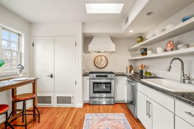 kitchen with a skylight, white cabinetry, appliances with stainless steel finishes, custom range hood, and sink