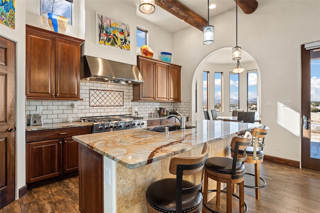 kitchen with dark hardwood / wood-style flooring, wall chimney exhaust hood, beam ceiling, light stone countertops, and an island with sink