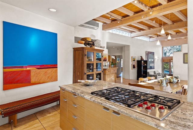 kitchen featuring stainless steel gas cooktop, a wall mounted air conditioner, light tile patterned floors, beamed ceiling, and light stone countertops