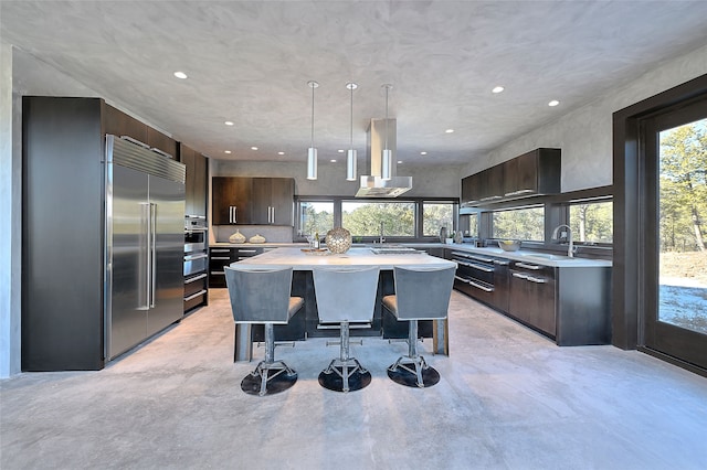 kitchen with stainless steel built in refrigerator, a center island, dark brown cabinets, a healthy amount of sunlight, and wall chimney range hood