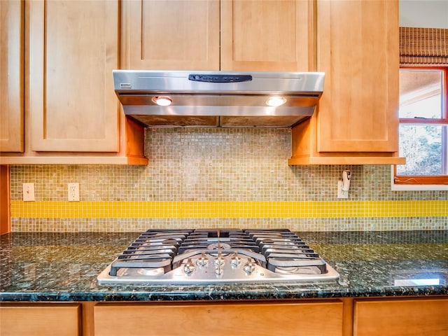 kitchen featuring stainless steel gas stovetop, dark stone countertops, backsplash, and range hood