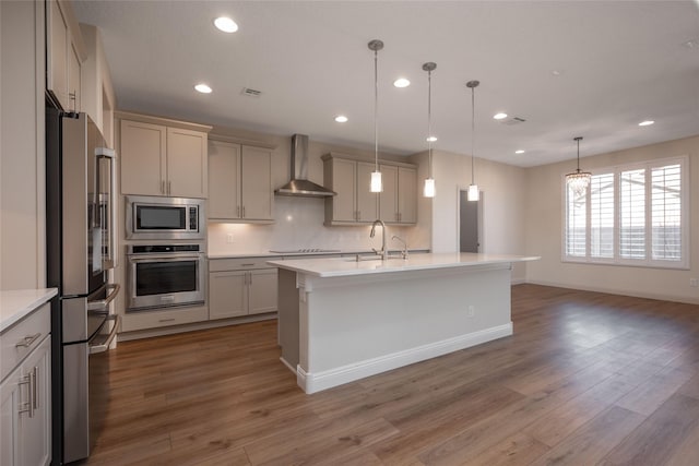 kitchen featuring appliances with stainless steel finishes, pendant lighting, sink, a kitchen island with sink, and wall chimney range hood