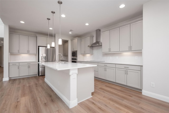 kitchen featuring wall chimney range hood, light hardwood / wood-style flooring, hanging light fixtures, stainless steel appliances, and an island with sink