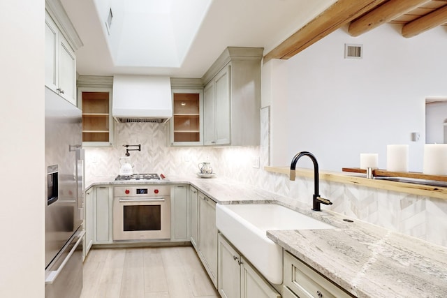 kitchen featuring appliances with stainless steel finishes, backsplash, beam ceiling, light stone countertops, and custom exhaust hood