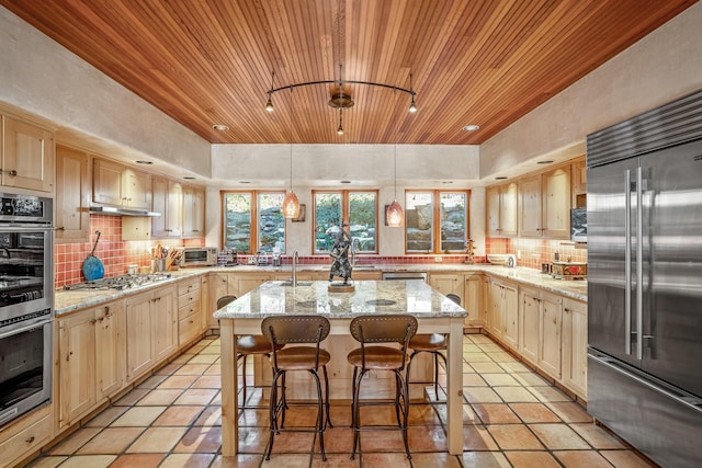 kitchen with light brown cabinetry, light stone countertops, stainless steel appliances, and an island with sink