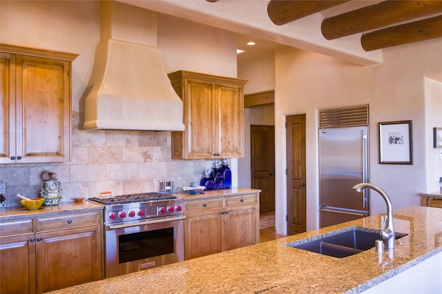 kitchen with sink, premium range hood, high end appliances, light stone counters, and decorative backsplash