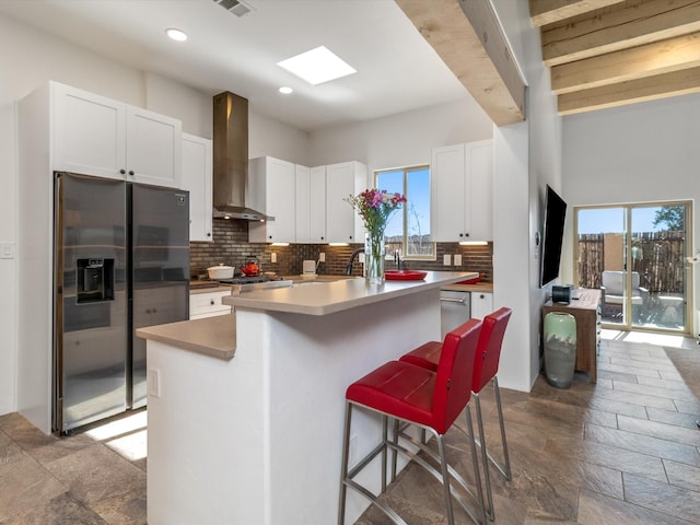 kitchen with a breakfast bar, an island with sink, white cabinets, stainless steel fridge, and wall chimney exhaust hood