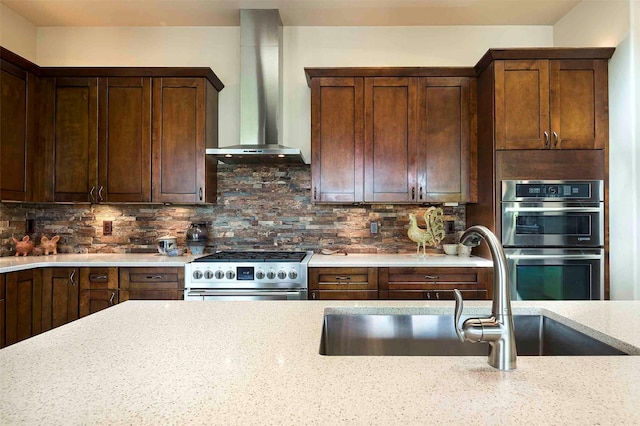 kitchen with backsplash, appliances with stainless steel finishes, a sink, wall chimney range hood, and light stone countertops