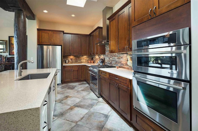 kitchen featuring light stone counters, a sink, appliances with stainless steel finishes, wall chimney range hood, and tasteful backsplash