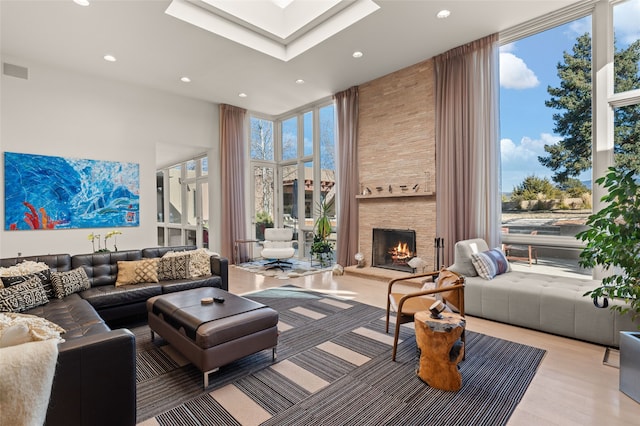 living room with a wall of windows, a skylight, a high ceiling, a stone fireplace, and light wood-type flooring