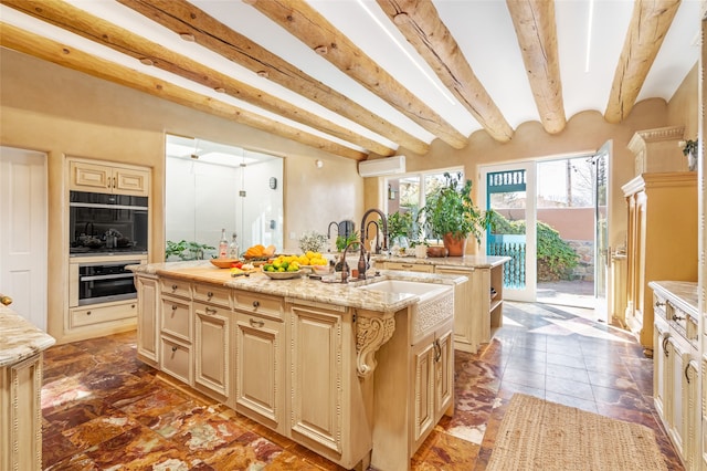 kitchen featuring sink, beam ceiling, light stone counters, an island with sink, and an AC wall unit