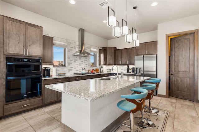 kitchen with sink, black double oven, wall chimney range hood, an island with sink, and light stone countertops