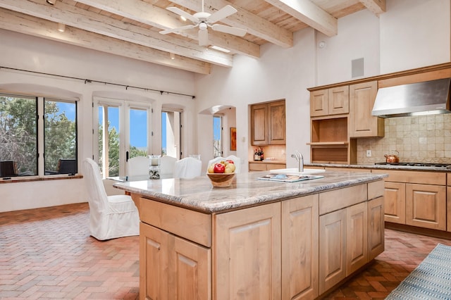 kitchen with wall chimney range hood, a center island with sink, stainless steel gas cooktop, and light brown cabinetry