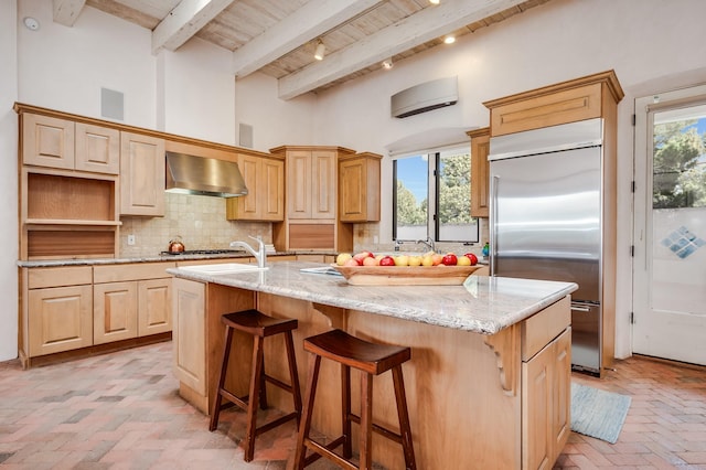 kitchen with tasteful backsplash, exhaust hood, a kitchen island with sink, light brown cabinetry, and built in fridge