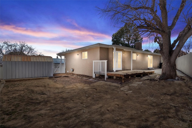 back of property at dusk with an outdoor structure, fence, stucco siding, a storage unit, and a chimney