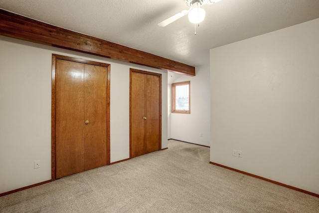 unfurnished bedroom featuring two closets, light colored carpet, a textured ceiling, beamed ceiling, and baseboards