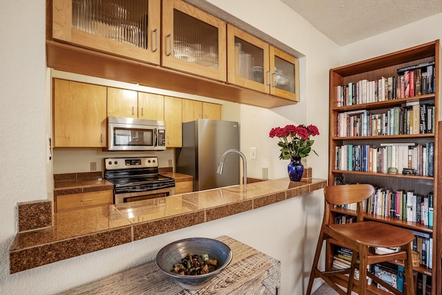 kitchen with stainless steel appliances, tile counters, glass insert cabinets, light brown cabinets, and a textured ceiling