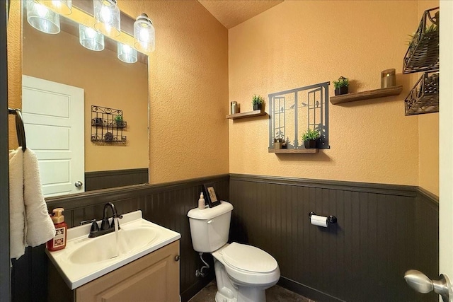 half bathroom with toilet, a textured wall, vanity, and wainscoting