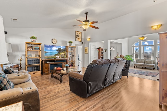 living area with a warm lit fireplace, high vaulted ceiling, visible vents, a ceiling fan, and light wood finished floors