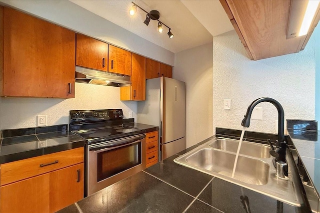 kitchen featuring dark countertops, appliances with stainless steel finishes, brown cabinets, under cabinet range hood, and a sink