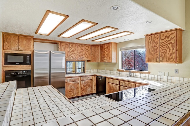 kitchen with a textured ceiling, a sink, tile counters, brown cabinets, and black appliances