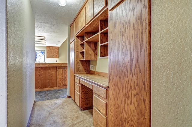 kitchen with a textured ceiling, a textured wall, built in desk, and light colored carpet