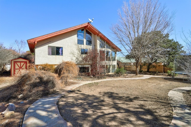 rear view of property featuring an outbuilding, a trampoline, fence, a storage unit, and stucco siding