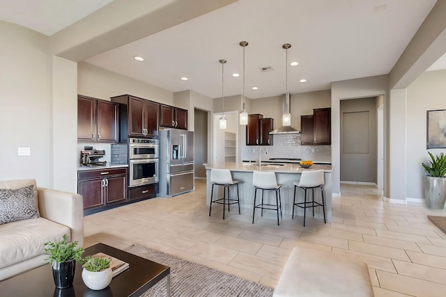 kitchen with wall chimney exhaust hood, stainless steel appliances, light countertops, a kitchen bar, and backsplash