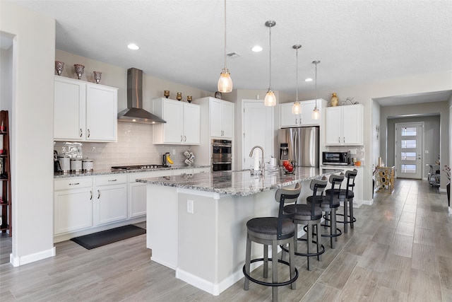 kitchen featuring a kitchen island with sink, stainless steel appliances, white cabinets, wall chimney range hood, and a kitchen bar