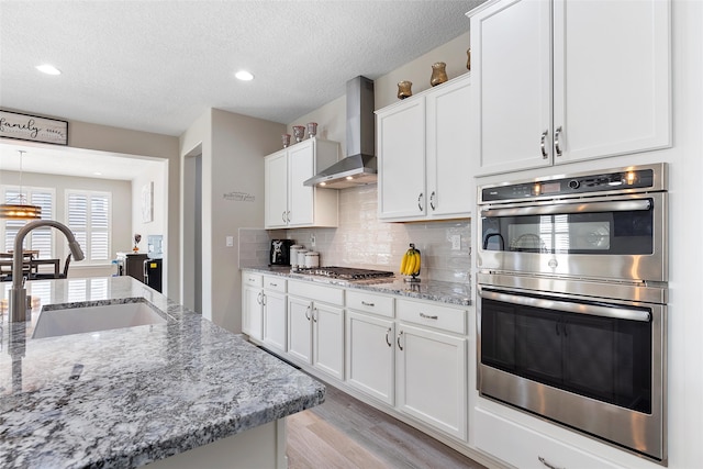 kitchen with stainless steel appliances, a sink, white cabinetry, wall chimney range hood, and backsplash