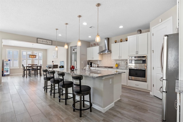 kitchen with wall chimney exhaust hood, backsplash, appliances with stainless steel finishes, a sink, and a kitchen breakfast bar